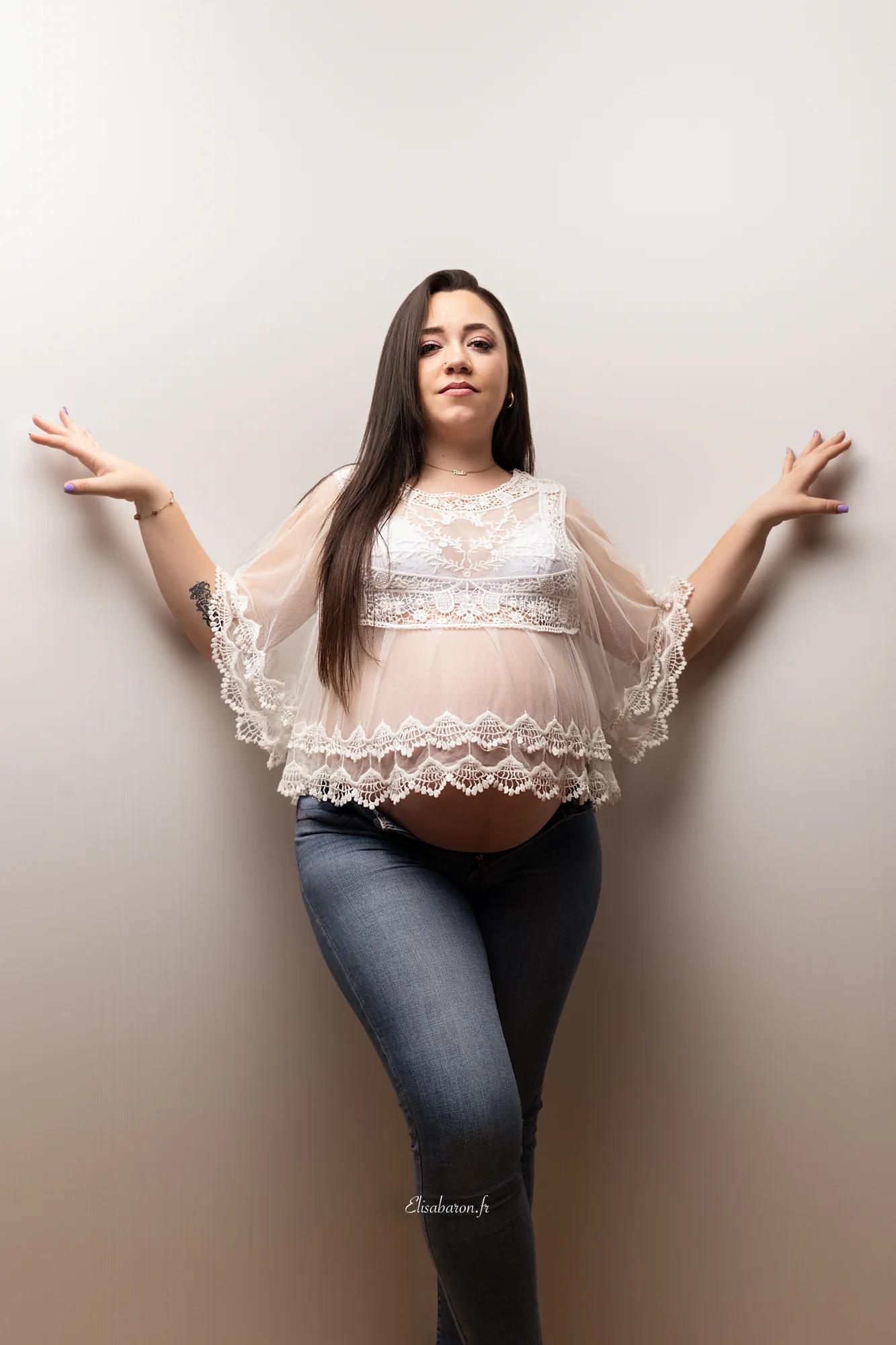 Femme enceinte en top blanc en dentelle et jean devant un fond clair au studio elisa baron près d'Angers
